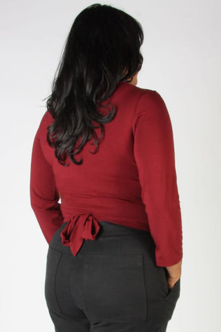 A woman viewed from the back wearing a long-sleeve red top with a tie detail at the waist and dark gray pants, against a plain white background.