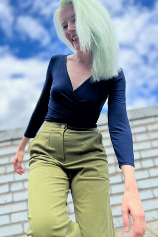 A woman with mint green hair wearing a navy blue long-sleeve wrap top and olive green trousers, photographed outdoors against a blue sky and brick wall background.