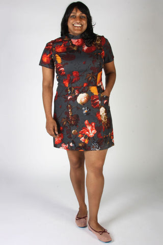 A woman wearing a short-sleeve dark gray sheath dress with red & orange floral print paired with pink flat shoes, against a plain white studio background.