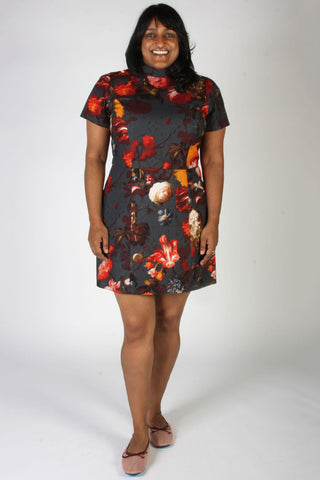 A woman wearing a short-sleeve dark gray sheath dress with red & orange floral print paired with pink flat shoes, against a plain white studio background.