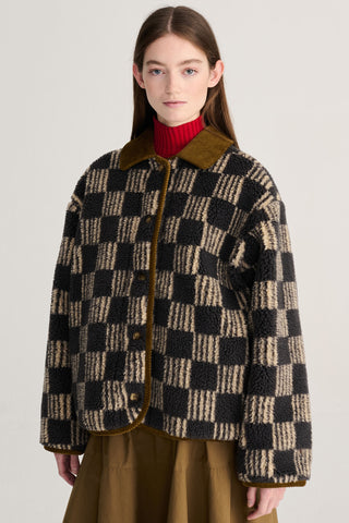 A model wearing a brown and beige checkered sherpa jacket with brown corduroy collar and trim, layered over a red turtleneck and brown skirt, against a light gray studio background.
