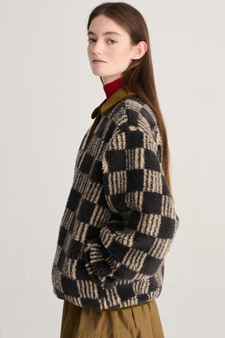 A woman wearing a brown and beige checkered sherpa jacket cardigan over a red turtleneck and olive green skirt, photographed against a plain light gray studio background.