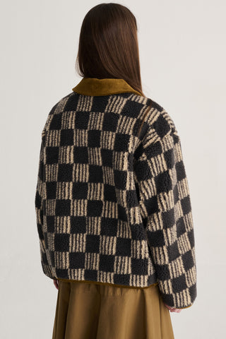 A person shown from the back wearing a brown and beige checkered sherpa jacket with brown collar, paired with a brown skirt, against a plain light gray background.