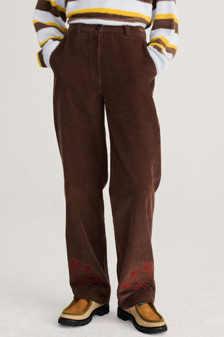 A person wearing brown corduroy trousers with red embroidered details near the hems, paired with a brown, white, and yellow striped sweater and tan loafers, shown from the chest down against a light gray studio background.