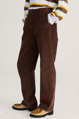A person wearing brown corduroy trousers with red embroidered details near the hems, paired with a brown, white, and yellow striped sweater and tan loafers, shown from the chest down against a light gray studio background.