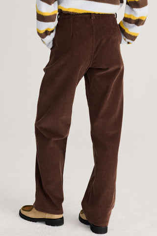 A person wearing brown corduroy trousers with red embroidered details near the hems, paired with a brown, white, and yellow striped sweater and tan loafers, shown from the back, waist down against a light gray studio background.