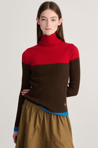 A model wearing a ribbed colorblock turtleneck sweater with a bright red yoke, dark brown sleeves and body, and a blue hem detail, paired with a brown skirt against a light gray studio background.