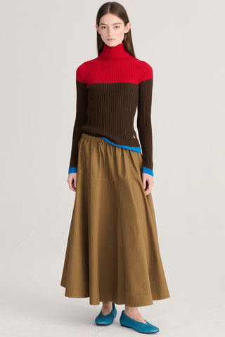 A model wearing a color-blocked ribbed turtleneck sweater in red, brown, and blue, paired with a brown midi skirt and blue flats, against a light gray studio background.