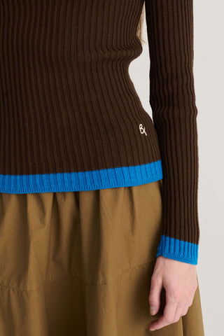 Close-up of a ribbed brown sweater with bright blue ribbed trim at the hem and cuffs, worn with a brown skirt. A small white embroidered logo is visible on the hem.