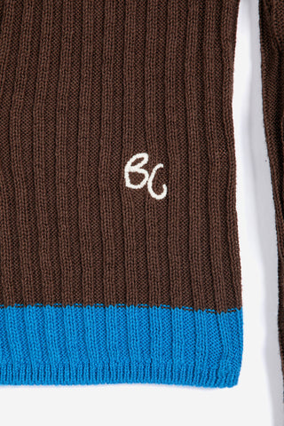 Close-up of a ribbed knit garment in dark brown with a bright blue stripe at the hem and a small white embroidered monogram logo.