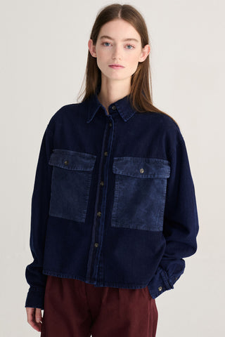 A model wearing a dark navy blue button-up shirt jacket with contrasting textured chest pockets, paired with burgundy pants, against a plain light gray background.
