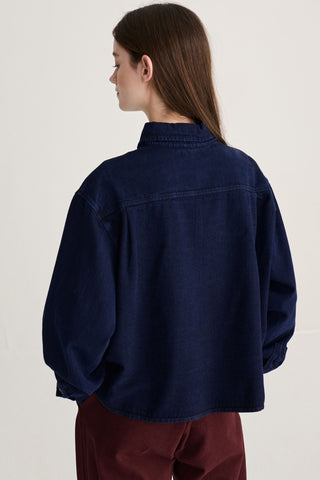 A woman shown from the back wearing an oversized dark navy blue denim shirt-jacket and burgundy pants, photographed against a plain off-white studio background.