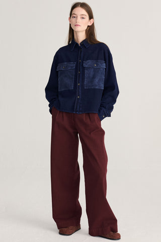 A model stands against a light gray studio background wearing a dark blue denim shirt with large corduroy pockets, paired with wide-leg burgundy trousers and brown slippers.