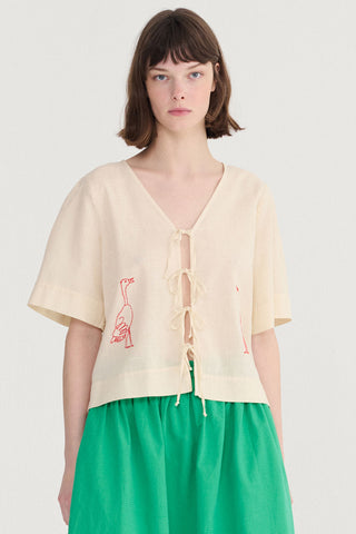 Woman wearing cream blouse with tie front and embroidered bird, with green skirt. 