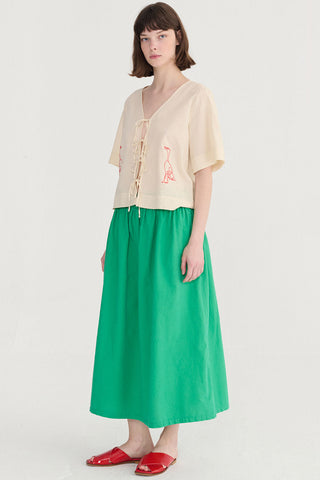 Woman wearing cream blouse with tie front and embroidered bird, with green skirt and red sandals. 