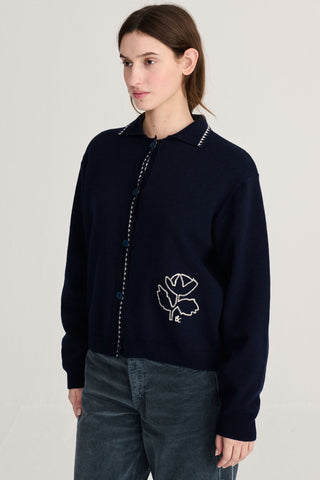 A woman wearing a navy blue knit cardigan with white embroidered details, paired with blue corduroy pants against a plain light gray studio background.