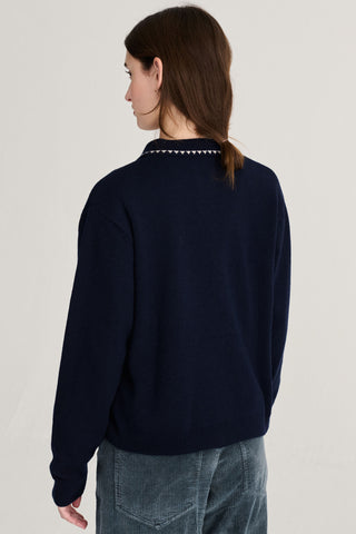 A woman shown from the back wearing a navy blue knit sweater with a white embroidery around the collar, paired with blue corduroy pants against a plain light gray studio background.