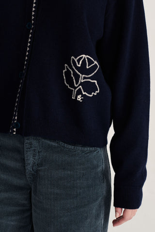 Close-up of a navy blue knit cardigan with a white embroidered flower motif, worn with blue corduroy trousers against a light neutral background.