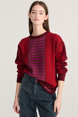 A model wearing a red and dark red striped knit sweater with a central magenta checkered panel, paired with dark blue cords, against a light gray studio background.