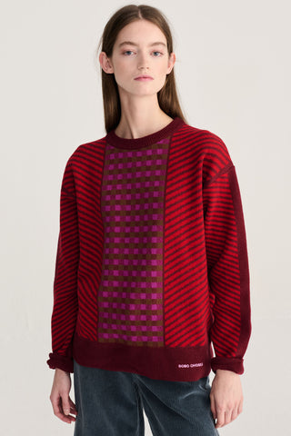 A model wearing a red and dark red striped knit sweater with a central magenta checkered panel, paired with dark blue cords, against a light gray studio background.
