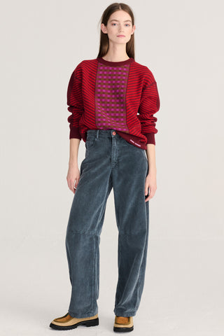 A model stands against a light gray studio background wearing a red and purple striped knit sweater with a purple grid pattern on the chest, dark blue corduroy pants, and tan loafers.