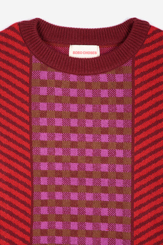 A close-up of a knit sweater by Bobo Choses, featuring a burgundy crew neck, red and burgundy diagonal striped sleeves, and a central brown and purple checkered pattern panel.