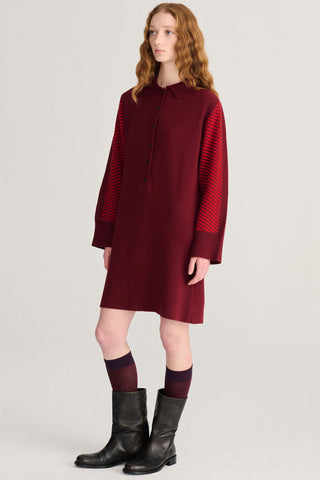A model wearing a long-sleeved burgundy dress with diagonal pattern on sleeves, paired with dark socks and black leather boots, photographed against a light gray studio background.