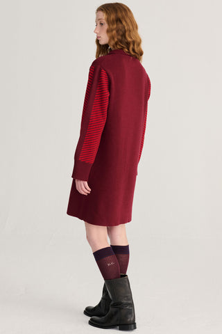 A model is shown from the back wearing a burgundy knit dress with red striped knit sleeves, paired with dark socks and black boots, against a plain light gray studio background.