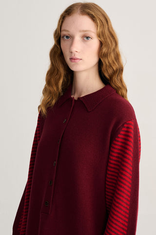 A model with long wavy red hair wearing a burgundy knit dress with red and burgundy striped sleeves, photographed against a light gray studio background.