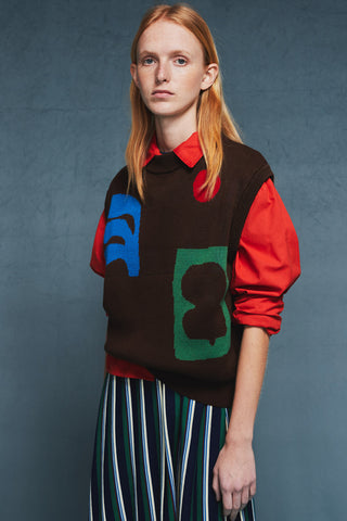 Woman in front of blue background wearing red collared shirt with brown knit vest over top paired with striped skirt. 