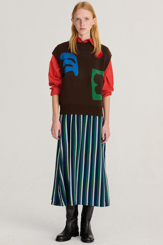 A model in front of light gray studio background wearing red shirt under a brown sweater vest with large blue and green graphic shapes on the chest, paired with a long striped skirt and black leather boots. 
