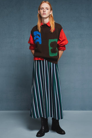 Woman in front of blue background wearing red collared shirt with brown knit vest over top paired with striped skirt and black leather boots. 