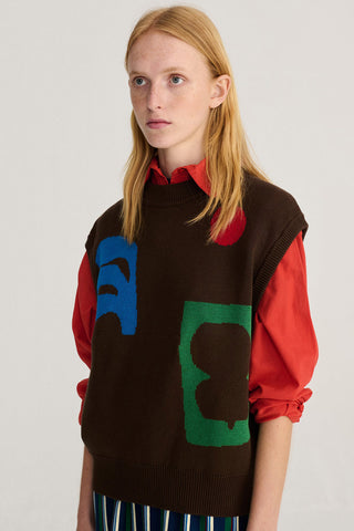 A model wearing a brown knit sweater vest with large blue and green graphic patches over a red collared shirt, photographed against a light gray studio background.