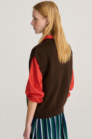 A woman shown from the back wearing a brown knit sweater vest over a bright red long-sleeve blouse, against a plain light gray background.