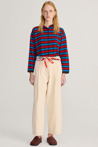 A model stands against a light gray studio background wearing a long-sleeve striped cardigan with red, blue, and black horizontal stripes, paired with wide-leg cream pants and brown slip-on shoes.
