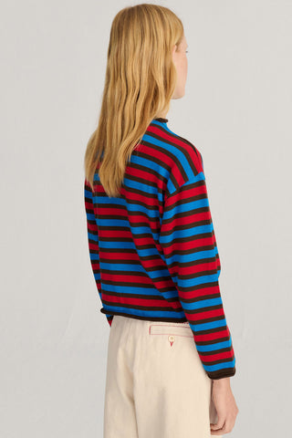 A model in a light gray studio shown from the back wearing a long-sleeve knit cardigan with horizontal red, blue, and navy stripes, paired with cream trousers.