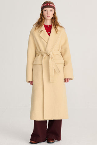 A model wearing a long, light yellow wool-blend coat with a matching belt, styled over a red top and dark burgundy wide-leg trousers, against a plain light gray studio background.