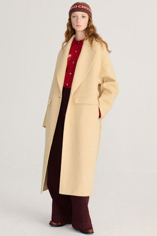 A model wearing a long light yellow wool coat over a red button-up shirt, dark burgundy trousers, and a brown beanie, standing against a light gray studio background.