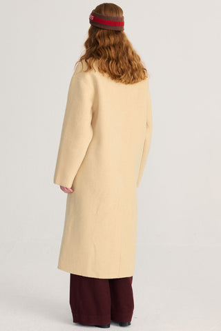 A person shown from the back wearing a long light yellow wool coat, wide-leg burgundy pants, photographed against a plain light gray studio background.