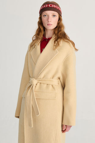 A model wearing a long, light yellow wool coat with a matching tie belt, a red collared shirt underneath, and a brown knit beanie, set against a plain light gray background.