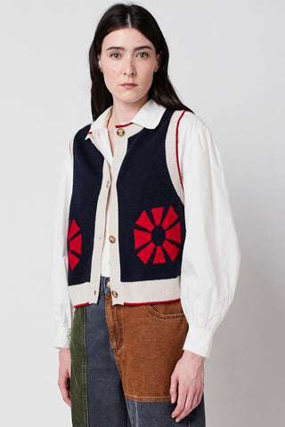 Woman wearing navy sleeveless button up knit vest with red geometric flower and cream trim by Bobo Choses, over white shirt and patchwork denim. 