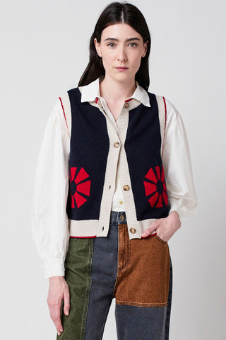 Woman wearing navy sleeveless button up knit vest with red geometric flower and cream trim by Bobo Choses, over white shirt and patchwork denim. 