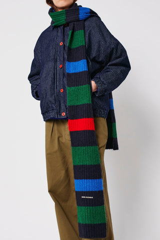 Woman wearing pinstripe denim jacket, olive pants, and multi colour stripe scarf by Bobo Choses. 