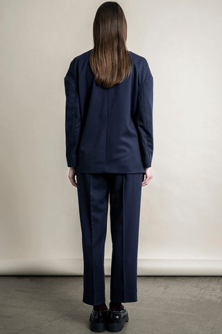 Back view of woman standing in front of beige background, wearing navy Regent Blazer and matching pants by Bodybag. 
