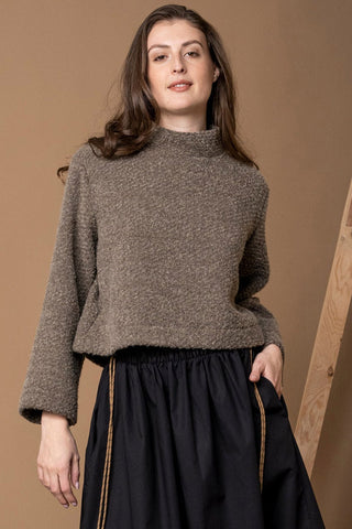 Woman standing in front of light brown wall wearing textured brown sweatshirt with high neckline, paired with pleated black skirt. 