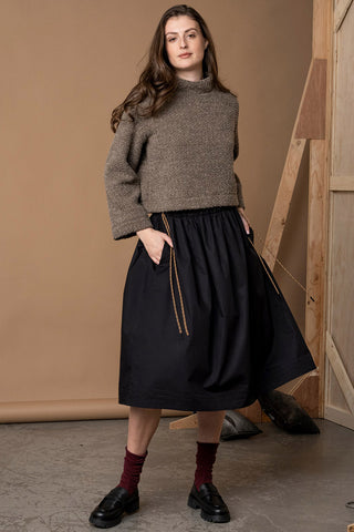 Woman standing in front of light brown wall wearing textured brown sweatshirt with high neckline, paired with pleated black skirt, burgundy socks and black loafers. 
