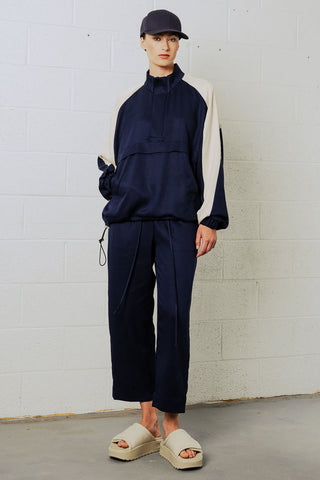 Woman in front of white brick wall wearing navy blue baseball cap, navy and cream anorak and draw-string pants. 