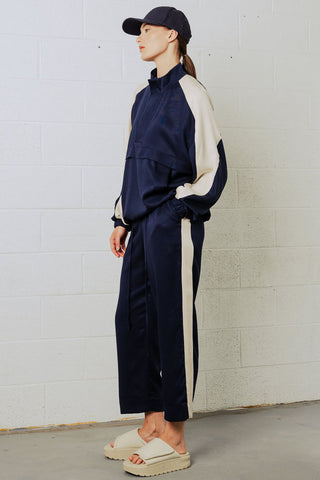 Woman in front of white brick wall wearing navy blue baseball cap, navy and cream anorak and draw-string pants with cream tuxedo stripe. 