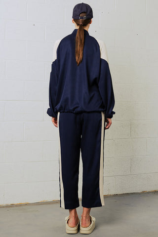 Back view of woman in front of white brick wall wearing navy blue baseball cap, navy anorak and navy pants with cream tuxedo stripe. 