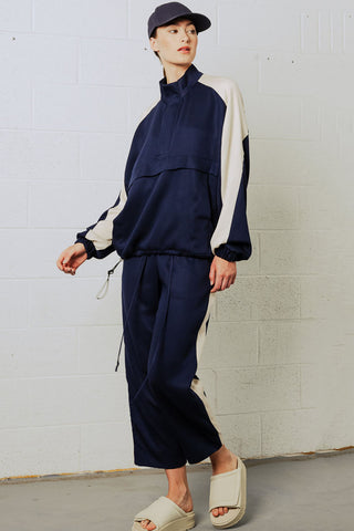 Woman in front of white brick wall wearing navy blue baseball cap, navy and cream jacket and draw-string pants with white tuxedo stripe. 